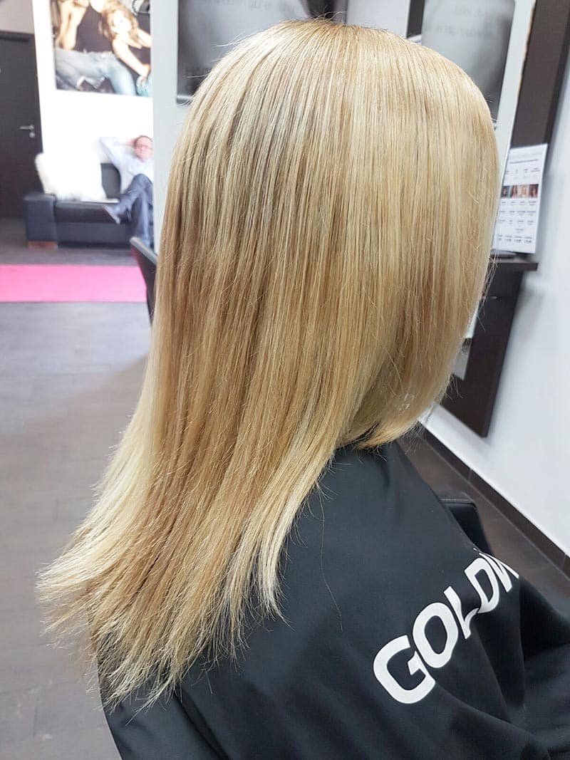 Glattes, hellblondes Haar reicht über die Schultern und fällt in gleichmäßigen Strähnen über einen schwarzen Friseurumhang.